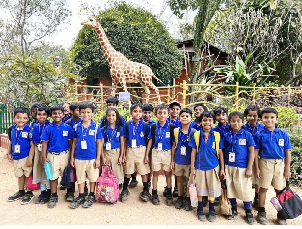 A group of smiling Guru Dronacharya Pathshala students in uniform on a school field trip to the zoo, posing in front of a giraffe statue.