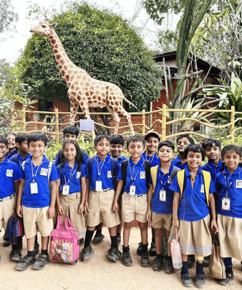A group of smiling Guru Dronacharya Pathshala students in uniform on a school field trip to the zoo, posing in front of a giraffe statue.